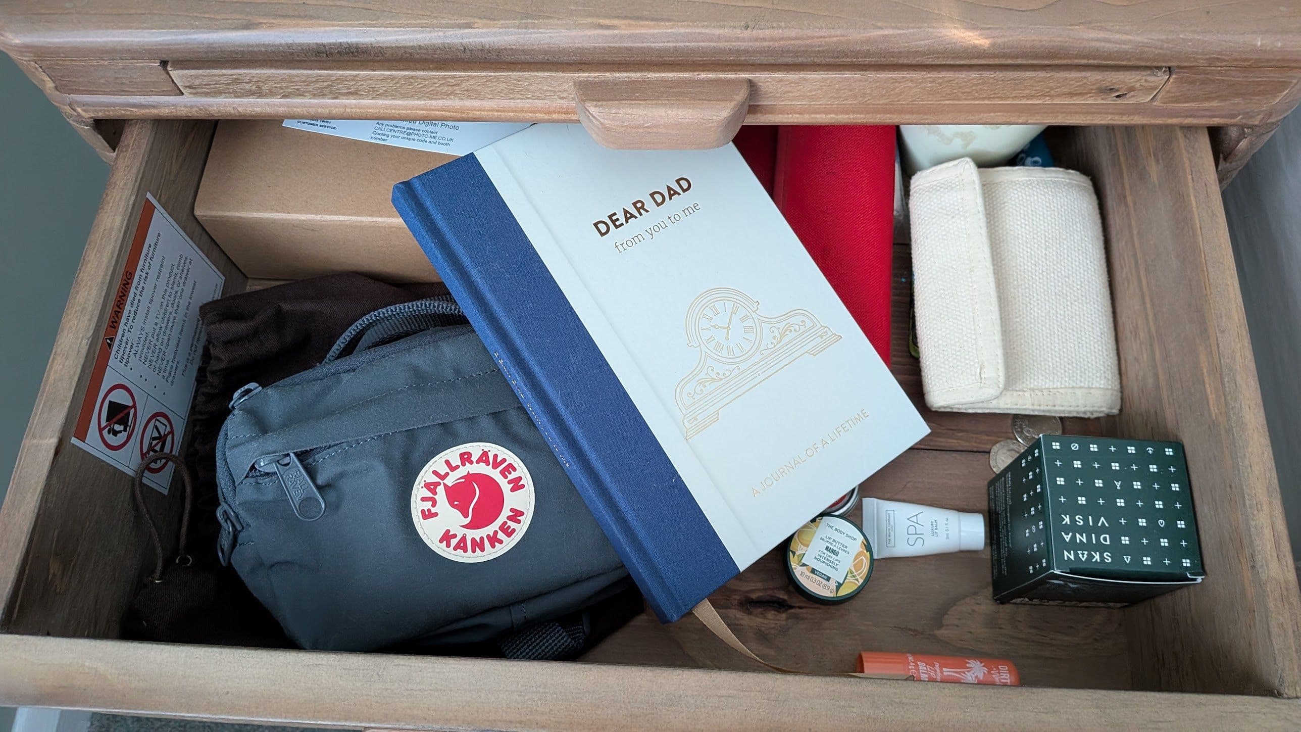 A bedside drawer pulled open, with a cream and blue hardback journal, Dear Dad: from you to me, lying on top of a grey bag and other everyday items.