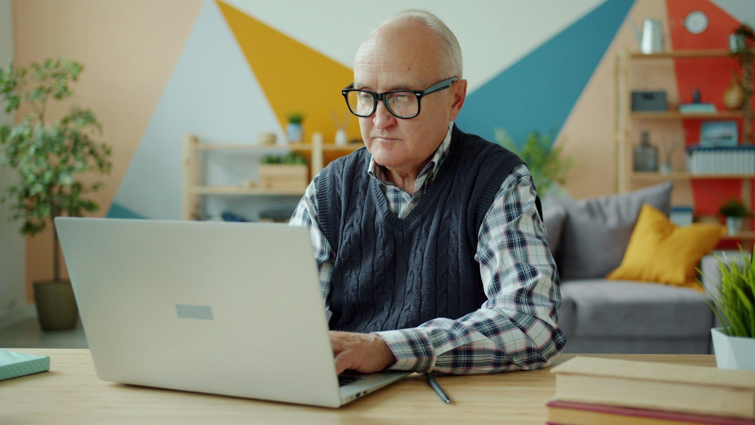 An older man with glasses sitting at a desk, typing on a laptop.